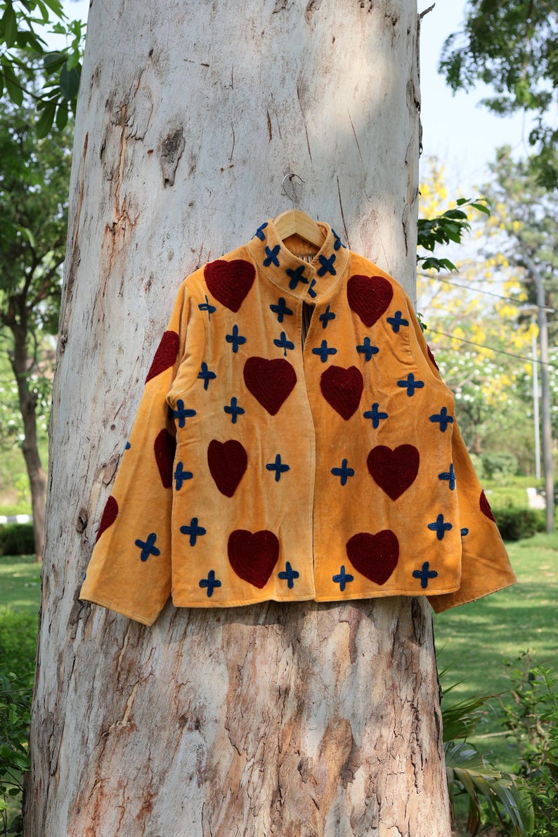 Puede incluir: Chaqueta color mostaza con cuello alto, adornada con grandes corazones burdeos y detalles de cruces azul marino. La chaqueta est&aacute; colgada en una percha de madera contra un tronco de &aacute;rbol, mostrando un dise&ntilde;o &uacute;nico y artesanal.