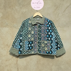 May include: A patchwork jacket with a variety of patterns in shades of green, blue, and beige. The jacket features a collared neckline, two front pockets, and striped cuffs. The jacket is hanging on a wooden hanger.
