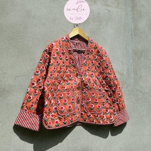 Könnte beinhalten: Gesteppte orangefarbene Jacke mit floralem Muster, zwei Taschen und gestreiftem Futter. Die Jacke hat einen V-Ausschnitt und ist geknöpft. Die Manschetten sind gestreift. Die Jacke hängt an einem Holzbügel.