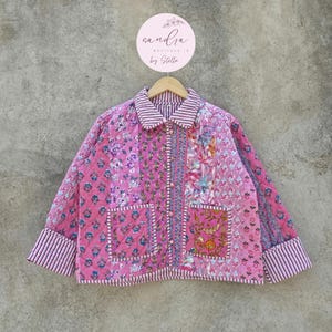 Pode incluir: Casaco acolchoado rosa com estampas florais e listradas. O casaco tem gola, fecho de botões e dois bolsos frontais. As mangas e a gola são adornadas com listras rosa e brancas. O casaco está pendurado em um cabide de madeira.