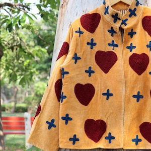 Puede incluir: Chaqueta de terciopelo amarillo mostaza con aplicaciones de corazones burdeos y cruces azules. La chaqueta est&aacute; colgada en una percha de madera, mostrando un dise&ntilde;o &uacute;nico y artesanal con detalles decorativos.