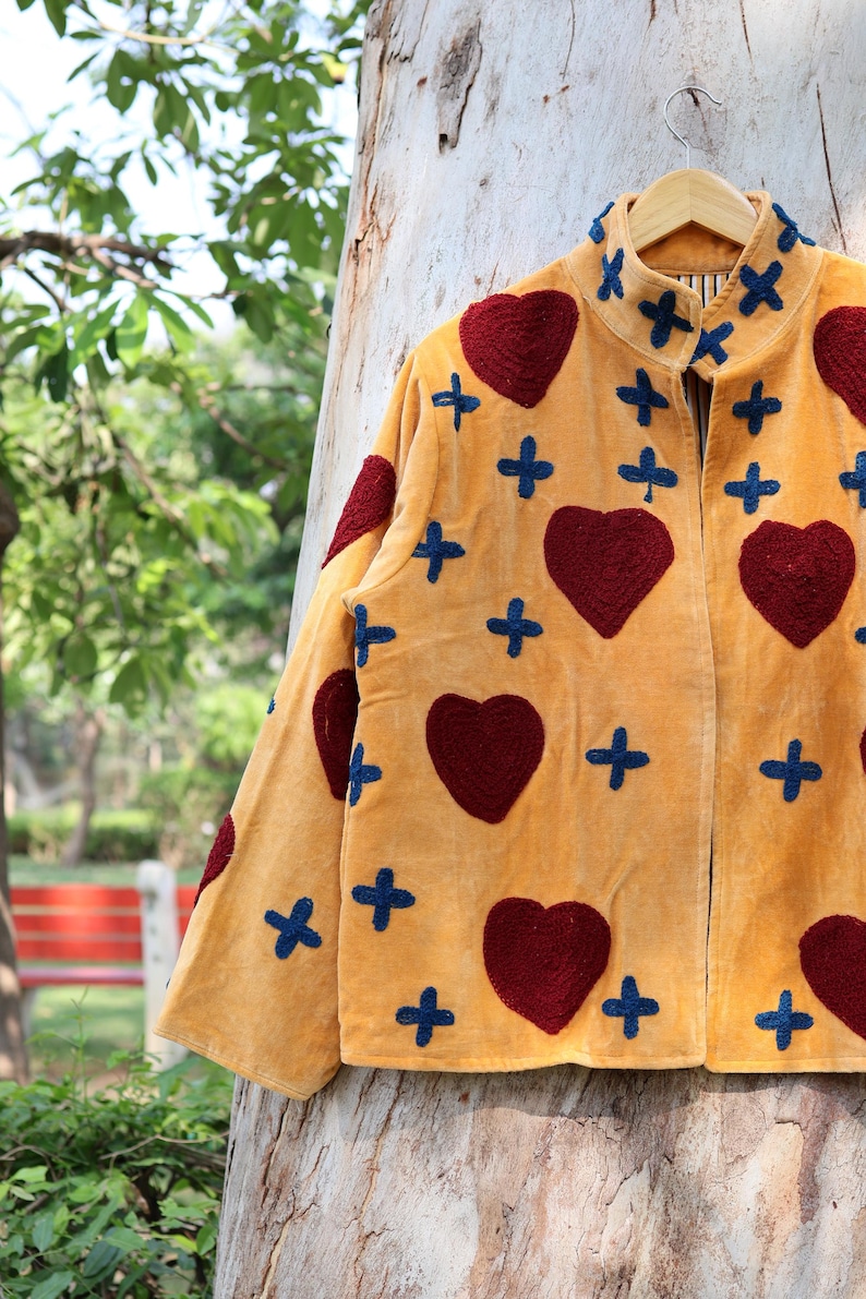 Puede incluir: Chaqueta de terciopelo amarillo mostaza con aplicaciones de corazones burdeos y cruces azules. La chaqueta est&aacute; colgada en una percha de madera, mostrando un dise&ntilde;o &uacute;nico y artesanal con detalles decorativos.