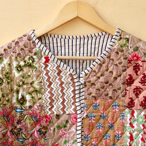 May include: A quilted jacket with a patchwork design featuring various floral patterns in shades of pink, brown, and green. The jacket has a striped trim and a button closure.