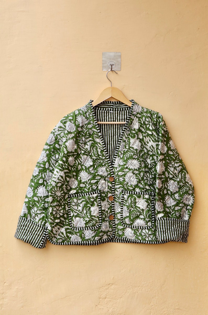 Puede incluir: Una chaqueta acolchada verde con estampado floral blanco y ribetes a rayas blanco y negro. La chaqueta tiene cuello en V y cierre de bot&oacute;n.