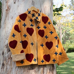 Puede incluir: Chaqueta amarillo mostaza con corazones burdeos y cruces azules. La chaqueta tiene cuello y pu&ntilde;os a rayas. Cuelga de una percha de madera contra un tronco de &aacute;rbol.