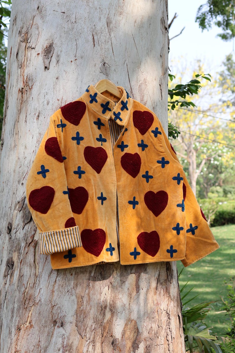 Puede incluir: Chaqueta amarillo mostaza con corazones burdeos y cruces azules. La chaqueta tiene cuello y pu&ntilde;os a rayas. Cuelga de una percha de madera contra un tronco de &aacute;rbol.