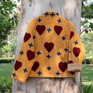 Puede incluir: Chaqueta amarillo mostaza con corazones burdeos y cruces azules. La chaqueta tiene cuello y pu&ntilde;os a rayas. Cuelga de una percha de madera contra un tronco de &aacute;rbol.