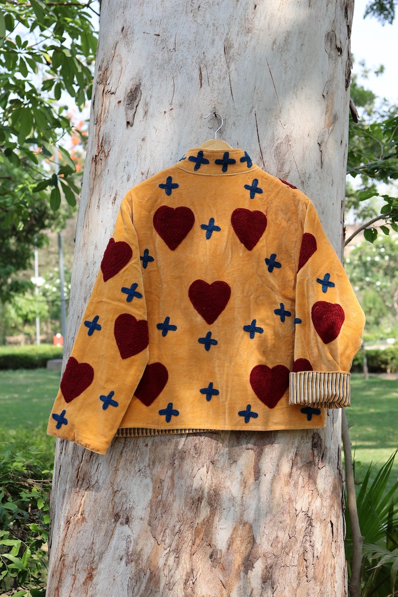 Puede incluir: Chaqueta amarillo mostaza con corazones burdeos y cruces azules. La chaqueta tiene cuello y pu&ntilde;os a rayas. Cuelga de una percha de madera contra un tronco de &aacute;rbol.