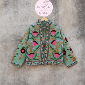 May include: A mint green embroidered jacket with a floral pattern. The jacket has a mandarin collar and a front zipper closure. The embroidery is in shades of pink, orange, and green. The jacket is hanging on a hanger with a round sign that says "sandra boutique by stella".
