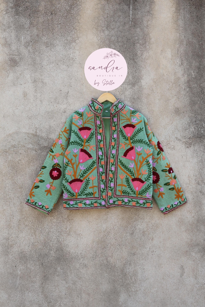 May include: A mint green embroidered jacket with a floral pattern. The jacket has a mandarin collar and a front zipper closure. The embroidery is in shades of pink, orange, and green. The jacket is hanging on a hanger with a round sign that says "sandra boutique by stella".