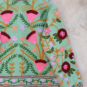 May include: A mint green jacket with floral embroidery in shades of pink, orange, and burgundy. The jacket has a mandarin collar and a button closure. The embroidery features a variety of flowers and leaves.
