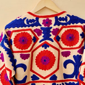 May include: A cream-colored jacket with intricate embroidery in shades of red, blue, and purple. The embroidery features floral and geometric patterns.