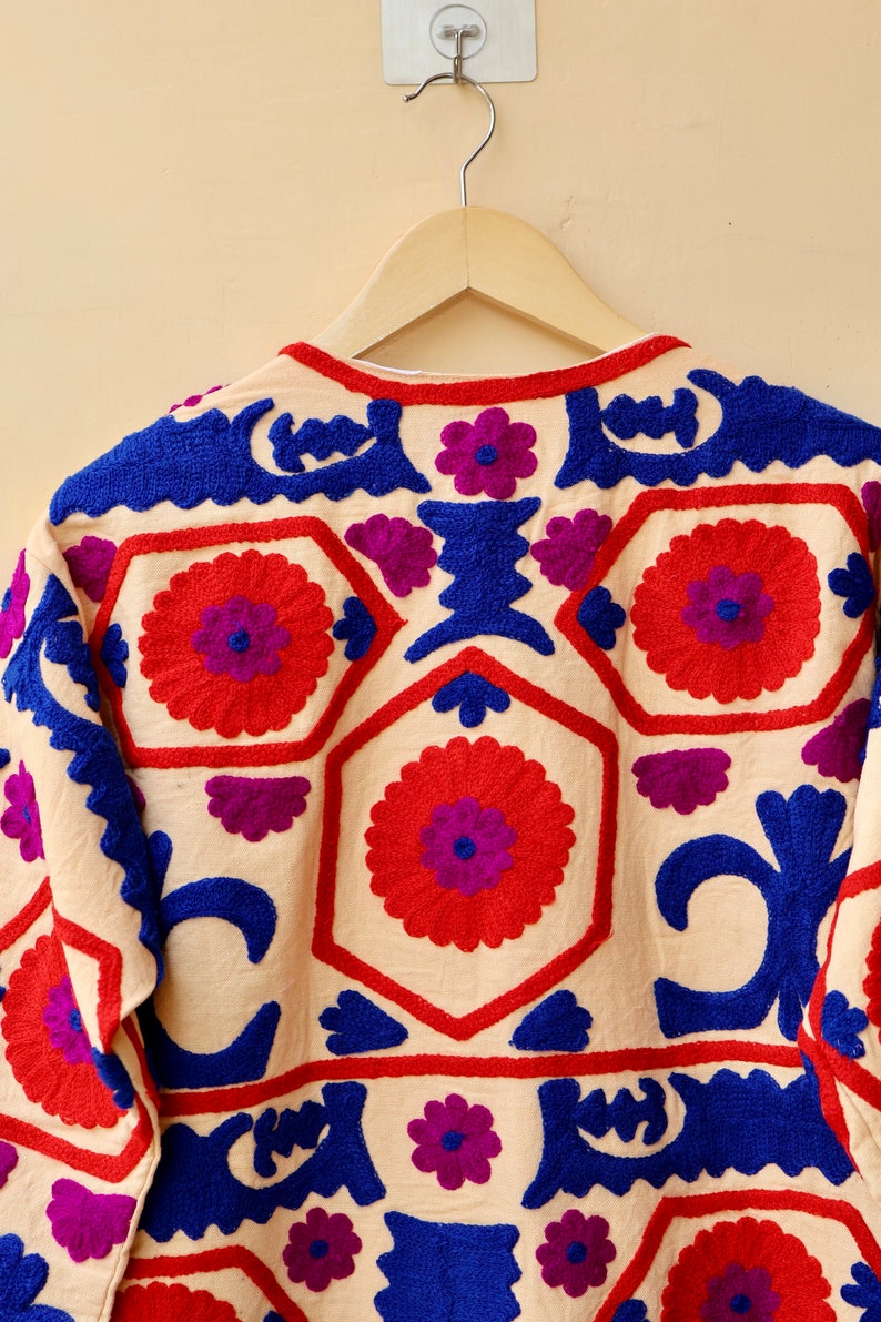 May include: A cream-colored jacket with intricate embroidery in shades of red, blue, and purple. The embroidery features floral and geometric patterns.