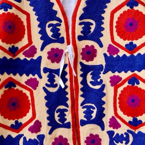 May include: A cream-colored fabric garment with a blue and red embroidered design. The garment has a V-neck and a tie closure. The embroidery features geometric shapes and floral motifs.