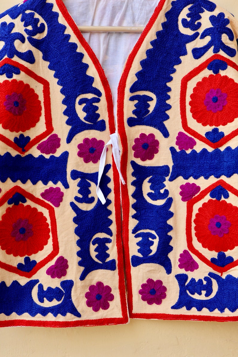May include: A cream-colored fabric garment with a blue and red embroidered design. The garment has a V-neck and a tie closure. The embroidery features geometric shapes and floral motifs.