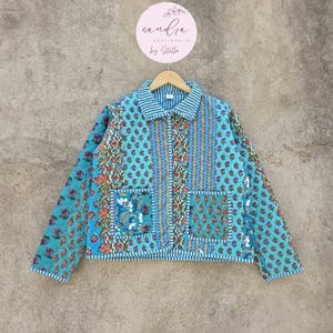 Puede incluir: Chaqueta acolchada turquesa con estampado floral y paisley. La chaqueta presenta cuello y puños a rayas, dos bolsillos delanteros y cierre de botones. La chaqueta está colgada de una percha de madera.