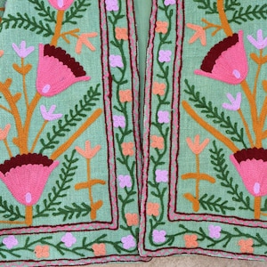 May include: A green linen jacket with colorful floral embroidery. The jacket has a mandarin collar and a front closure. The embroidery features pink, orange, purple, and green flowers.