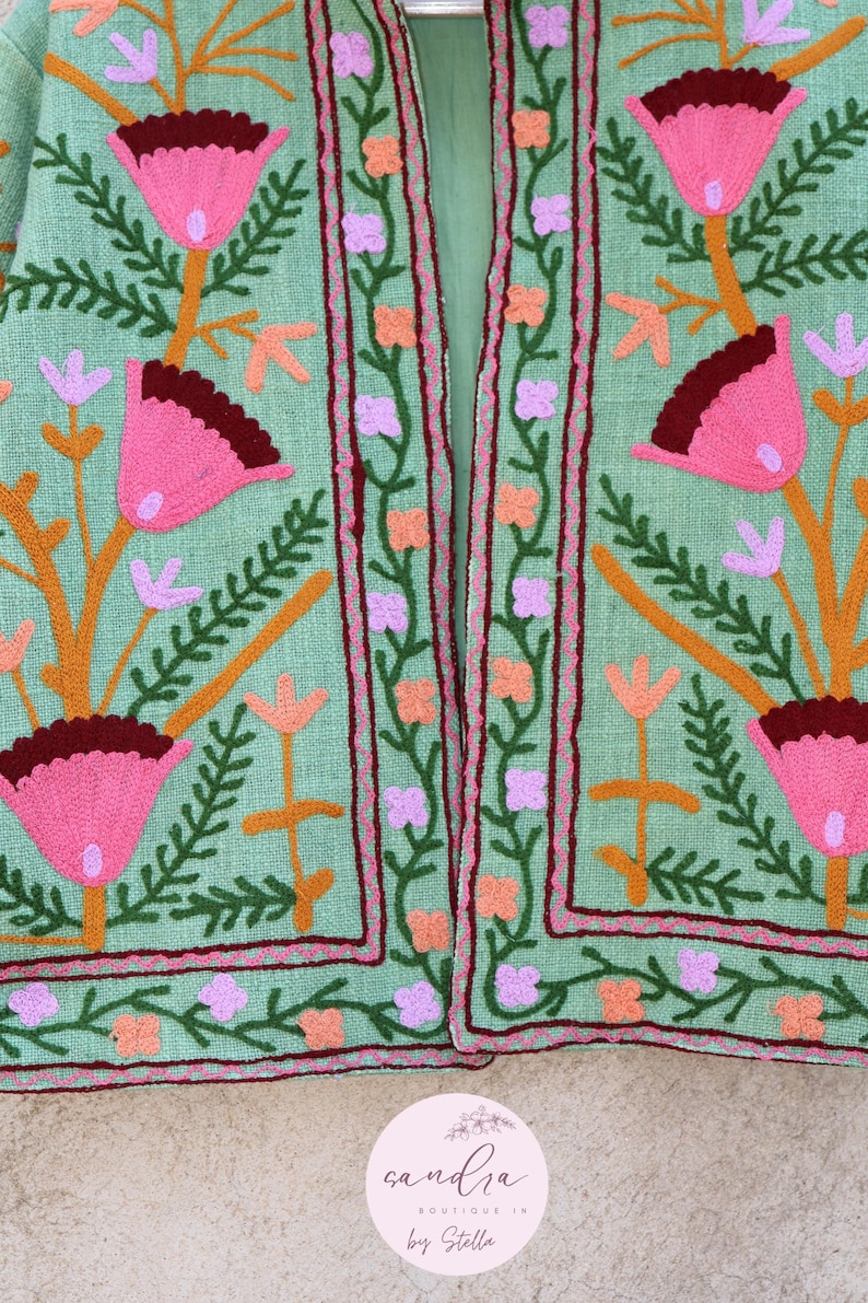May include: A green linen jacket with colorful floral embroidery. The jacket has a mandarin collar and a front closure. The embroidery features pink, orange, purple, and green flowers.