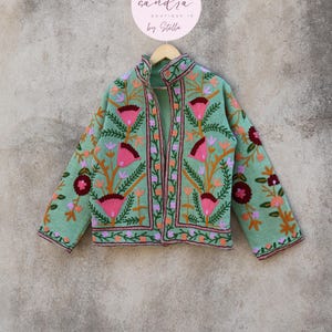May include: A mint green embroidered jacket with floral designs in pink, orange, and burgundy. The jacket has a mandarin collar and a front zipper closure. The text "candra BOUTIQUE IN by Stella" is on a pink circle.