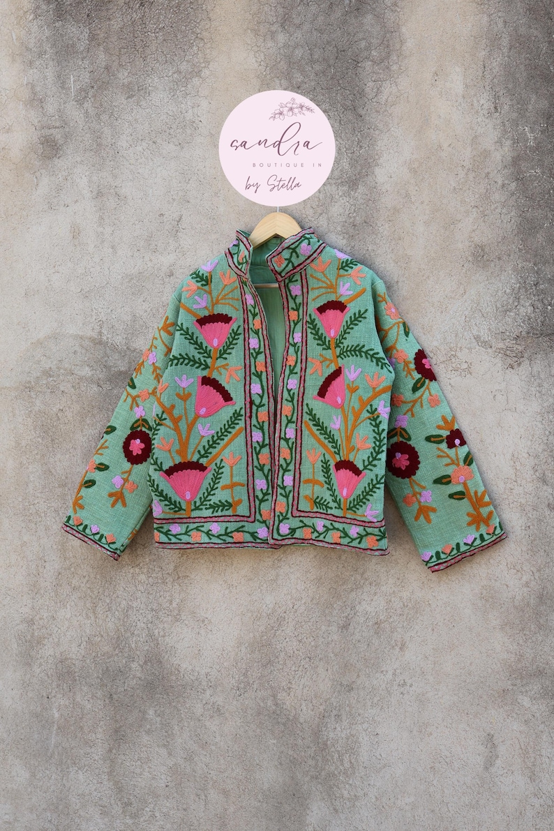 May include: A mint green embroidered jacket with floral designs in pink, orange, and burgundy. The jacket has a mandarin collar and a front zipper closure. The text "candra BOUTIQUE IN by Stella" is on a pink circle.