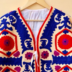 May include: A cream-colored jacket with blue, red, and purple embroidery. The jacket has a V-neck and a tie closure.