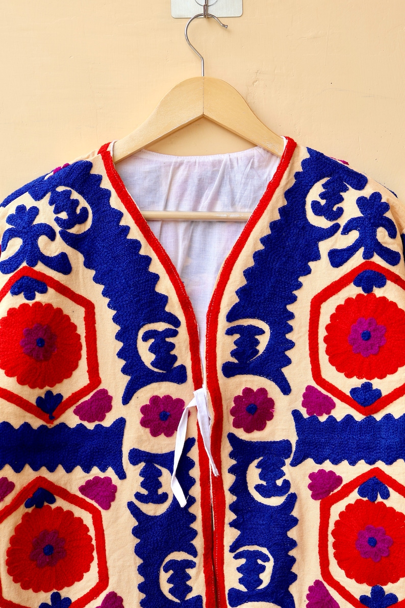 May include: A cream-colored jacket with blue, red, and purple embroidery. The jacket has a V-neck and a tie closure.