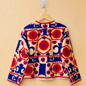 May include: A cream-colored jacket with long sleeves and a geometric pattern embroidered in red, blue, and purple.