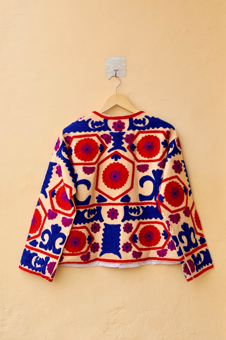May include: A cream-colored jacket with long sleeves and a geometric pattern embroidered in red, blue, and purple.