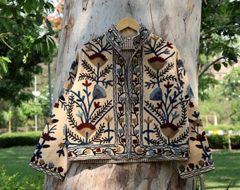 Puede incluir: Chaqueta beige con intrincados bordados florales en tonos azules, rojos y marrones. La chaqueta tiene un cuello alto y se muestra en una percha de madera. El bordado crea un patrón detallado en la superficie de la chaqueta.