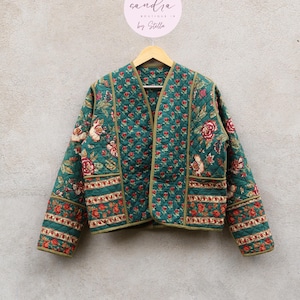 May include: Teal green quilted jacket with a floral design in red and cream. The jacket features a V-neck, long sleeves, and two pockets. It is displayed on a wooden hanger.