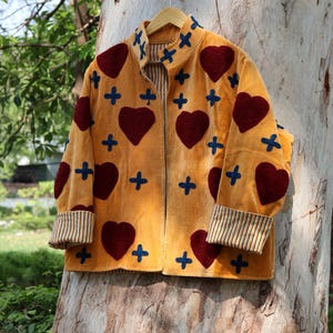 Puede incluir: Chaqueta amarillo mostaza con aplicaciones de coraz&oacute;n burdeos y cruz azul. La chaqueta tiene pu&ntilde;os a rayas y forro interior a rayas. La chaqueta est&aacute; colgada de una percha de madera contra un tronco de &aacute;rbol.