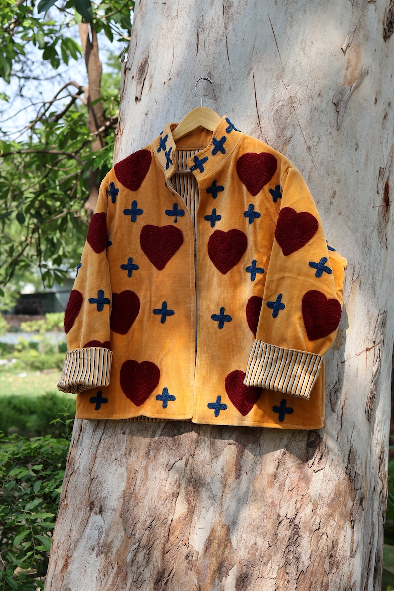 Puede incluir: Chaqueta amarillo mostaza con aplicaciones de coraz&oacute;n burdeos y cruz azul. La chaqueta tiene pu&ntilde;os a rayas y forro interior a rayas. La chaqueta est&aacute; colgada de una percha de madera contra un tronco de &aacute;rbol.