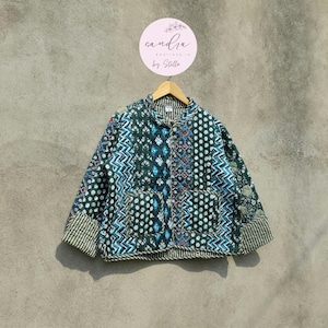 May include: A patterned quilted jacket in green, blue, and white, with a stand-up collar and two front pockets. The jacket is displayed on a wooden hanger. The brand name "Sandra Boutique by Stella" is visible in a circular logo.