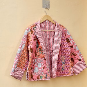 Op de afbeelding: Gewatteerde jas met een patchwork ontwerp in roze, perzik en wit. De jas heeft een gestreepte voering en bies, en een bloemen- en dierenprint patroon. Hij hangt aan een houten hanger.