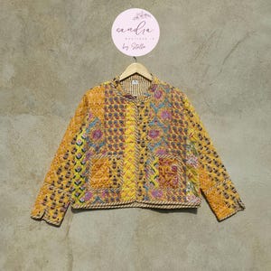 May include: A quilted jacket in shades of yellow, orange, and pink, featuring a floral and paisley pattern. The jacket has a round collar, button closure, and a decorative rope trim. The jacket is hanging on a wooden hanger.
