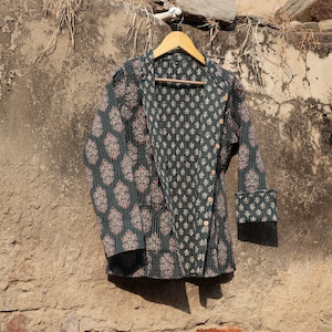 Puede incluir: Una chaqueta acolchada verde oscuro con estampado floral cuelga de una percha de madera contra una pared de ladrillo. La chaqueta tiene cierre de botón y mangas largas.