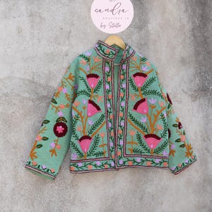 May include: A mint green embroidered jacket with floral patterns in pink, orange, and purple. The jacket has a mandarin collar and a front closure. The label on the hanger reads "sandra boutique in by Stella."