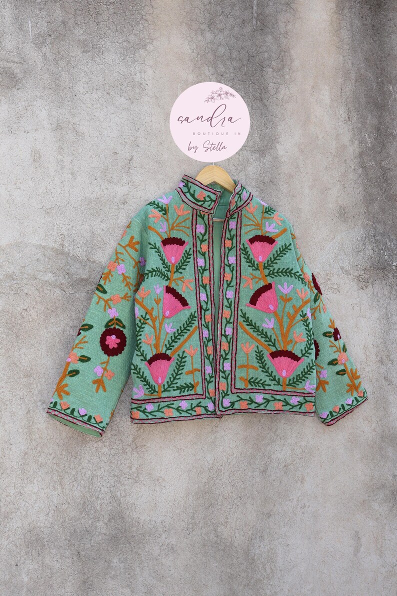 May include: A mint green embroidered jacket with floral patterns in pink, orange, and purple. The jacket has a mandarin collar and a front closure. The label on the hanger reads "sandra boutique in by Stella."