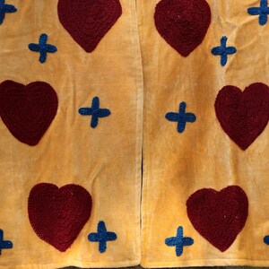 Puede incluir: Primer plano de una tela amarillo mostaza con un patr&oacute;n repetido de corazones burdeos y cruces azules. La tela parece suave y texturizada, posiblemente terciopelo o pana. El dise&ntilde;o es sim&eacute;trico.