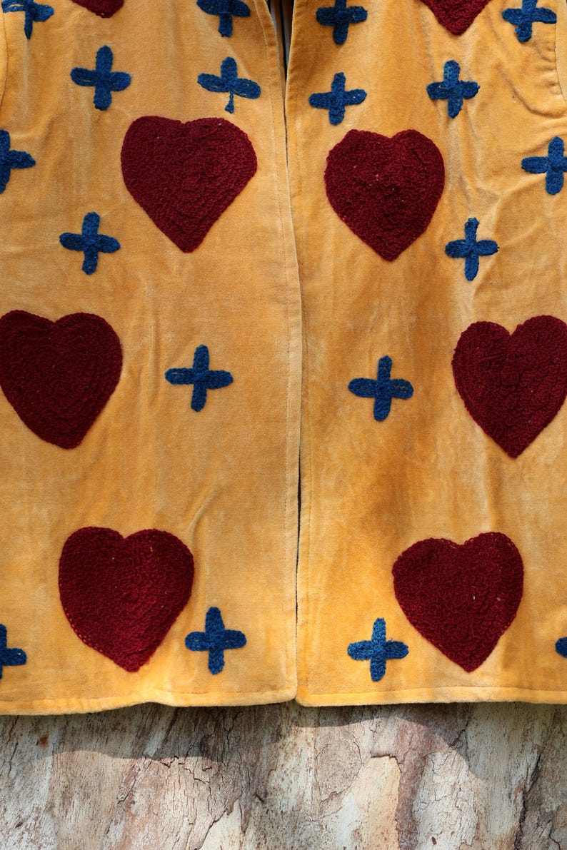 Puede incluir: Primer plano de una tela amarillo mostaza con un patr&oacute;n repetido de corazones burdeos y cruces azules. La tela parece suave y texturizada, posiblemente terciopelo o pana. El dise&ntilde;o es sim&eacute;trico.