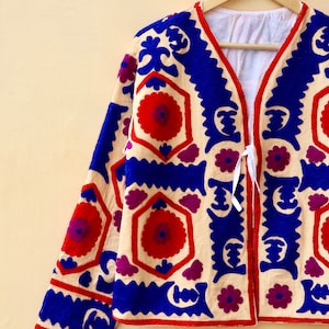 May include: A cream-colored jacket with a V-neck and long sleeves. The jacket is embroidered with a geometric pattern in shades of blue, red, and purple.