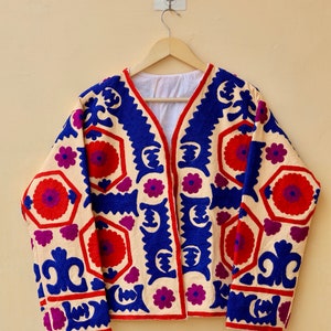 May include: A cream-colored jacket with a blue, red, and purple geometric pattern embroidered on the front and sleeves. The jacket has a V-neck and long sleeves.