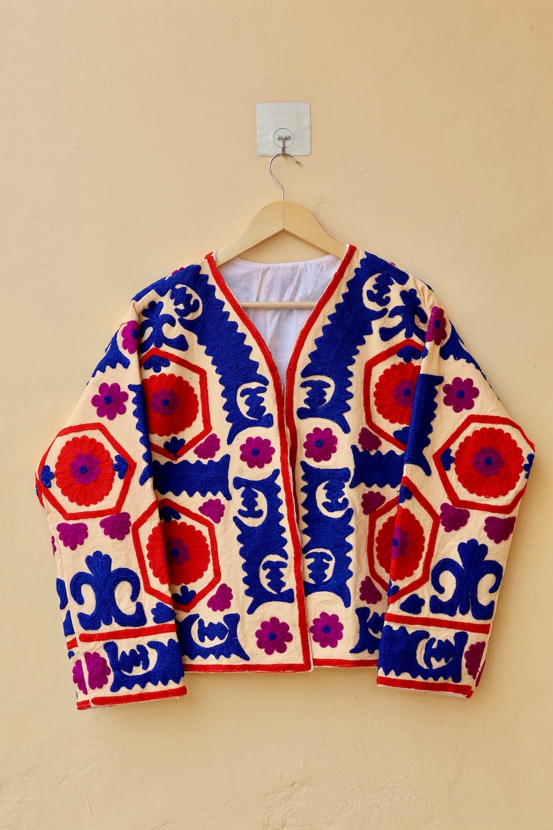 May include: A cream-colored jacket with a blue, red, and purple geometric pattern embroidered on the front and sleeves. The jacket has a V-neck and long sleeves.