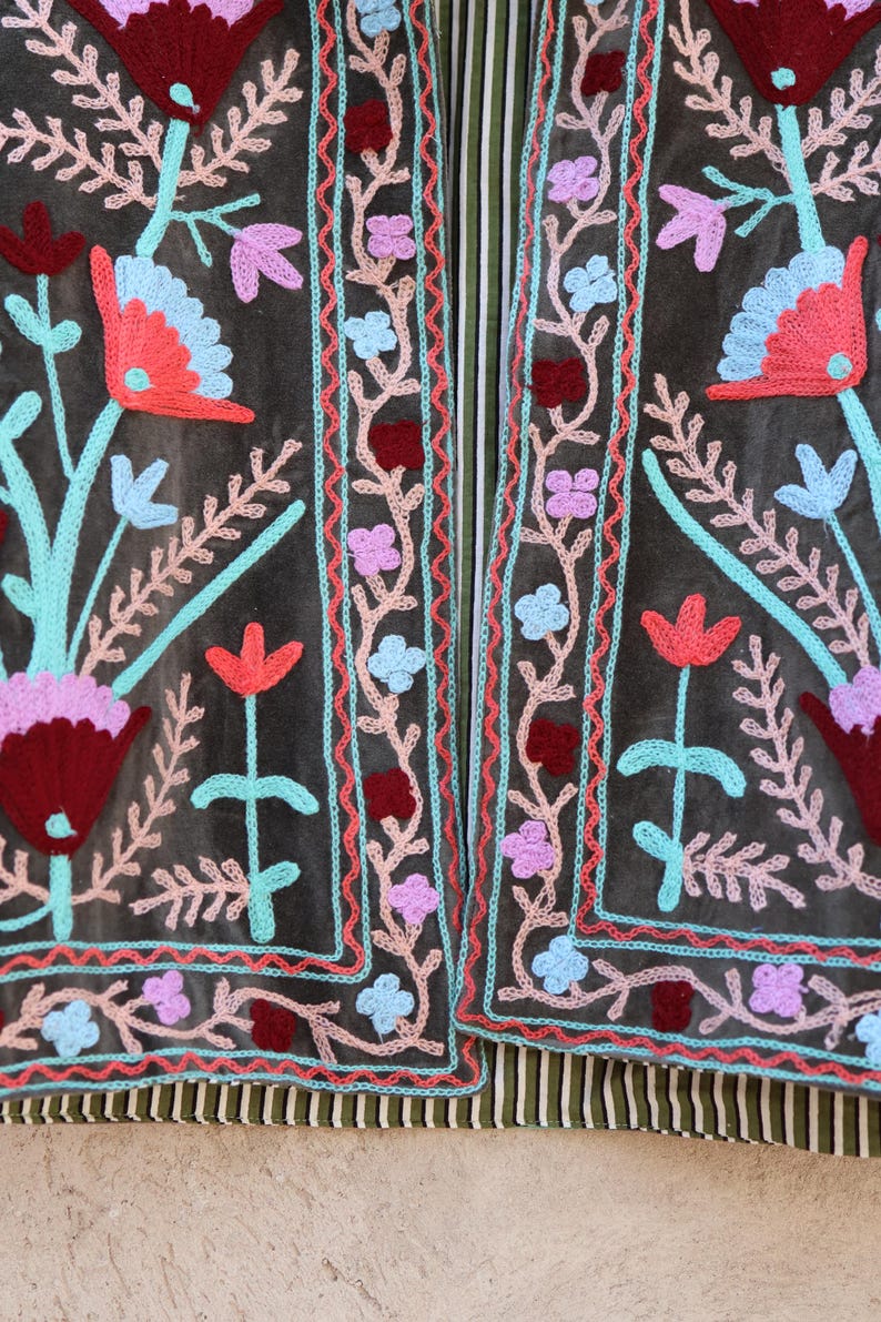 Peut inclure: Une veste en tissu gris fonc&eacute; avec un motif de broderie florale. La broderie pr&eacute;sente des fleurs et des feuilles roses, rouges, bleues et turquoise. La veste est doubl&eacute;e de rayures.