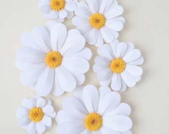 May include: A set of six white paper daisies with yellow centers. The daisies are arranged in a cluster on a white background.