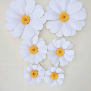 May include: Six white paper daisies with yellow centres of varying sizes. The daisies are arranged in a staggered pattern on a white background.