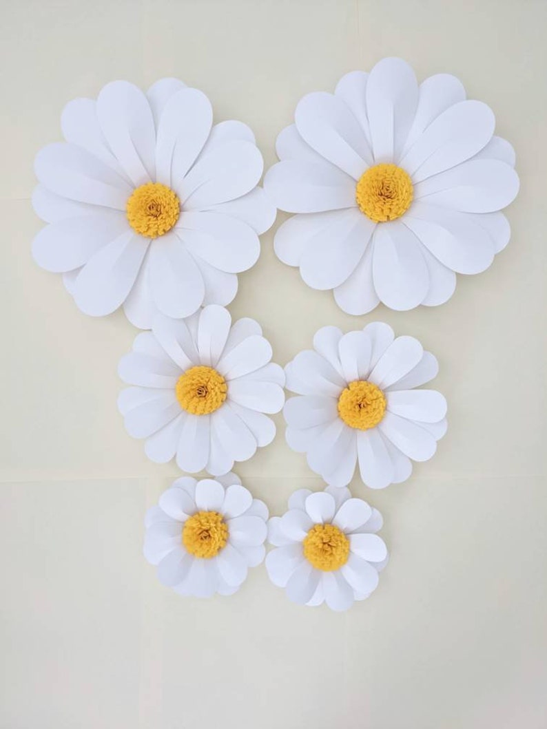 May include: Six white paper daisies with yellow centres of varying sizes. The daisies are arranged in a staggered pattern on a white background.
