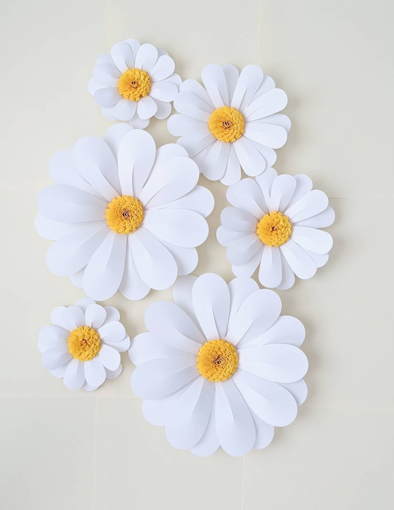 May include: A set of six white paper daisies with yellow centres. The daisies are arranged in a cluster on a white background.