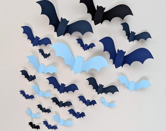 Op de afbeelding: Een verzameling papieren vleermuizen in verschillende tinten blauw en zwart, gerangschikt in een trapsgewijs patroon. De vleermuizen zijn van verschillende groottes, wat een dynamische en feestelijke Halloween-decoratie creëert.