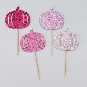 May include: Four pink and white glitter pumpkin cupcake toppers. The pumpkins are cut out of paper and have a sparkly finish. The toppers are attached to wooden sticks.